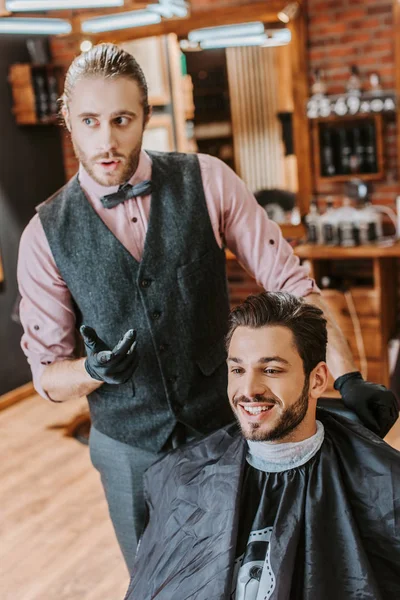 Guapo peluquero gesto cerca de feliz hombre en la barbería - foto de stock