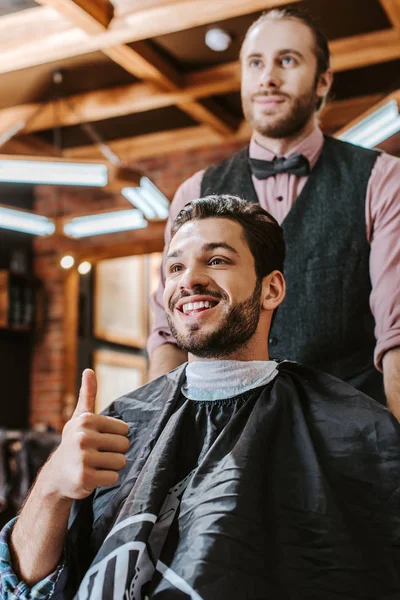 Enfoque selectivo de hombre feliz mostrando el pulgar hacia arriba cerca de barbero - foto de stock