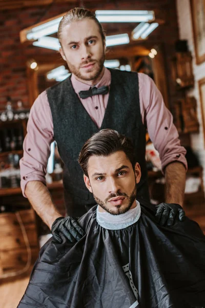 Barbeiro bonito em luvas de látex colocando as mãos sobre ombros de homem na barbearia — Fotografia de Stock