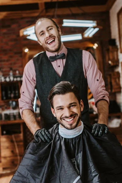 Guapo peluquero en guantes de látex poniendo las manos en los hombros de hombre feliz en la barbería - foto de stock