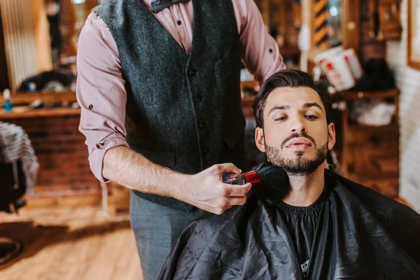 Peluquero celebración de cepillo cosmético cerca de la cara del hombre barbudo - foto de stock