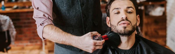 Tiro panorâmico de barbeiro segurando escova cosmética perto da face do homem barbudo — Fotografia de Stock