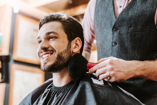 Peluquero celebración de cepillo cosmético cerca de la cara del hombre barbudo feliz - foto de stock