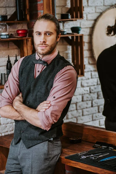 Handsome barber standing with crossed arms near scissors in barbershop — Stock Photo