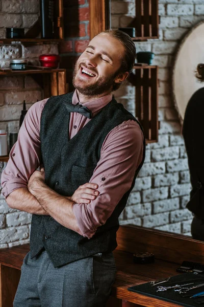 Cheerful barber standing with crossed arms and laughing in barbershop — Stock Photo