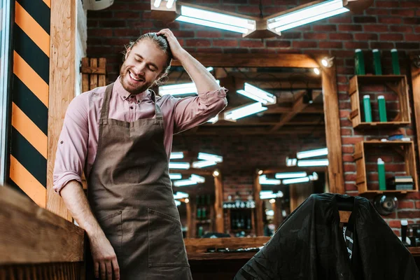 Selective focus of happy barber in apron touching hair in barbershop — Stock Photo