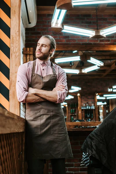 Selective focus of handsome barber in apron standing with crossed arms and looking away — Stock Photo