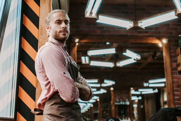 Beau coiffeur dans tablier debout avec les bras croisés et regardant la caméra — Photo de stock