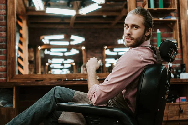 Beau coiffeur regardant la caméra tout en étant assis dans le fauteuil — Photo de stock