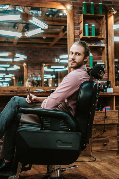 Beau barbier et barbu regardant la caméra tout en étant assis dans le fauteuil — Photo de stock