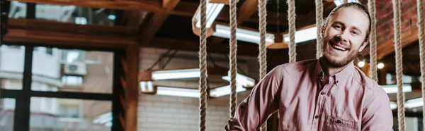 Panoramic shot of happy bearded man looking at camera and smiling in barbershop — Stock Photo