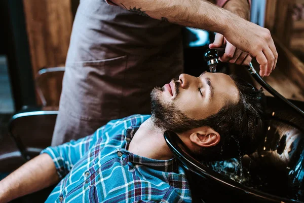 Peluquero tatuado lavando el cabello del hombre barbudo con los ojos cerrados - foto de stock