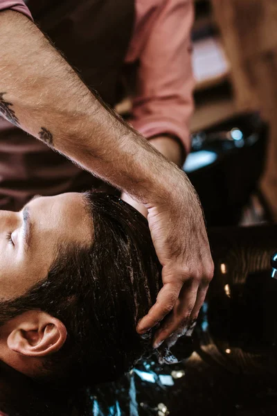 Обрезанный вид татуированного парикмахера, моющего волосы человека — стоковое фото