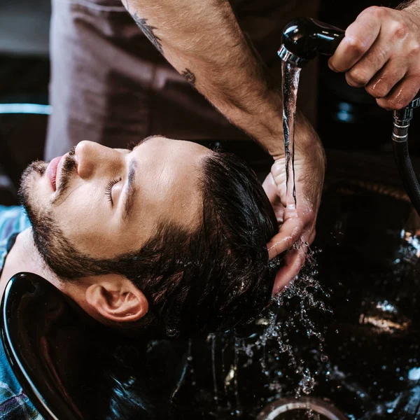 Tattooed barber washing wet hair of man in black sink — Stock Photo