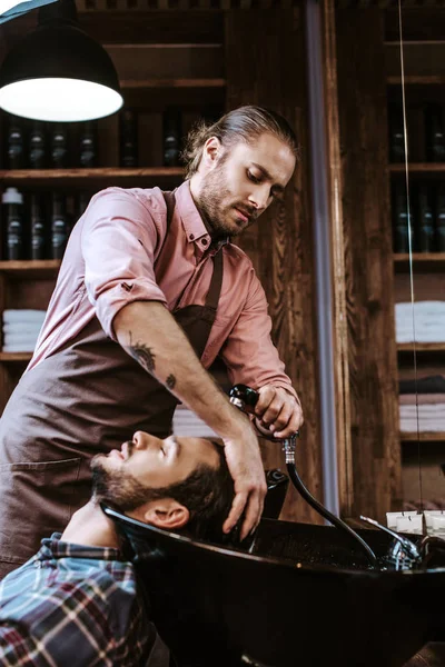 Guapo tatuado peluquero lavado húmedo cabello de hombre en negro fregadero - foto de stock
