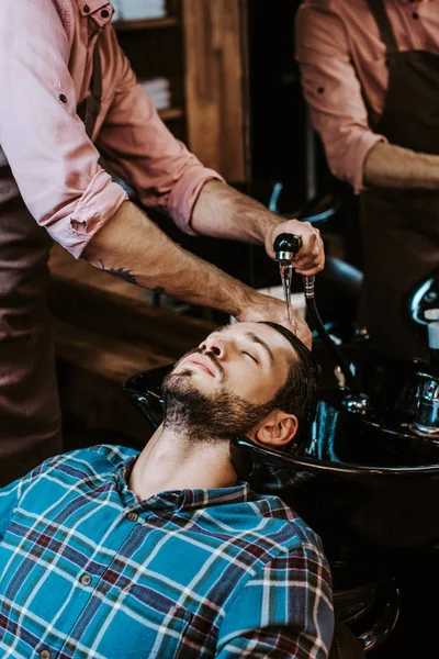 Татуйований перукар миє волосся людини в чорній раковині біля дзеркала — стокове фото
