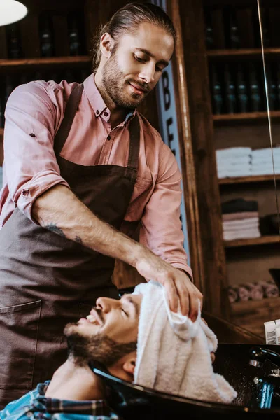 Selektiver Fokus eines hübschen Friseurs, der weißes Handtuch in der Nähe des Kopfes eines glücklichen Mannes hält — Stockfoto