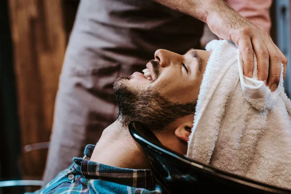 Barbero sosteniendo toalla blanca cerca de la cabeza del hombre feliz con los ojos cerrados - foto de stock