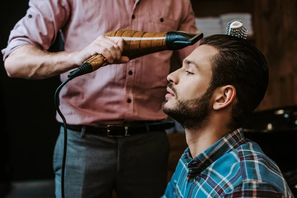 Парикмахер держит фен рядом с бородатым мужчиной в парикмахерской — стоковое фото