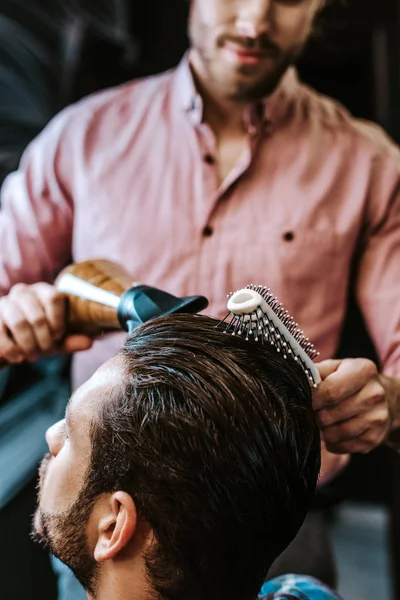 Selective focus of barber brushing hair and holding hair dryer near bearded man — Stock Photo