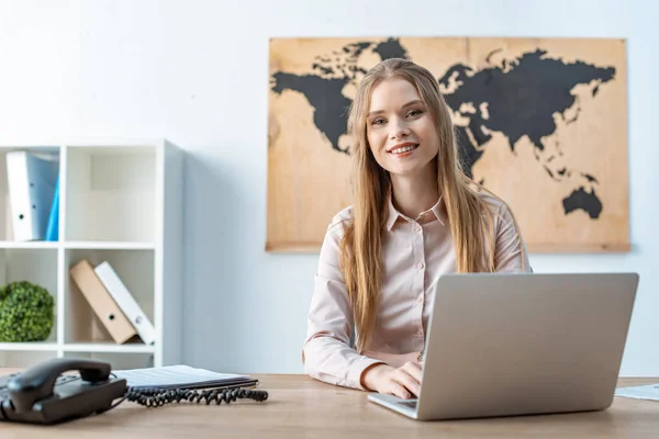 Attractive travel agent smiling at camera while using laptop — Stock Photo