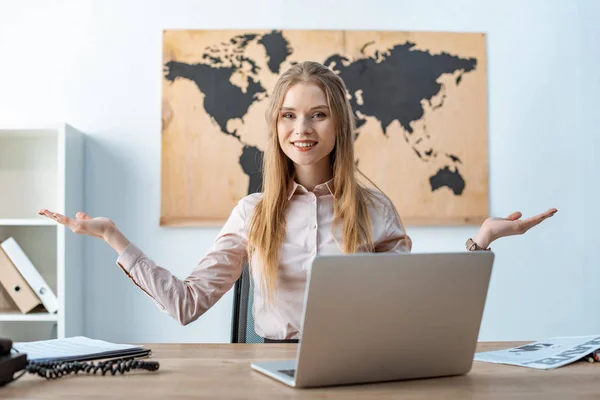 Smiling travel agent showing welcome gesture while sitting near laptop — Stock Photo