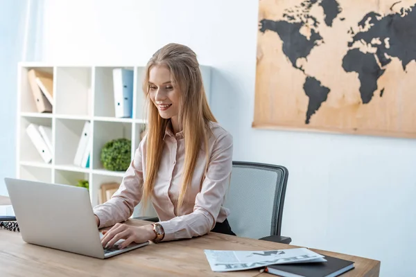 Agente de viajes sonriente utilizando el ordenador portátil mientras está sentado en el lugar de trabajo - foto de stock