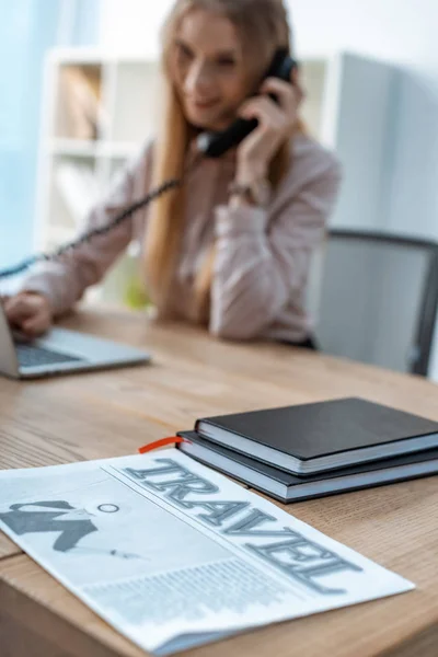 Selective focus of travel newspaper and notebooks on desk near travel agent talking on phone — Stock Photo