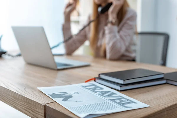 Selective focus of travel newspaper and notebooks on desk near travel agent talking on phone — Stock Photo