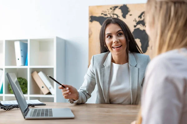 Agente de viajes sonriente hablando con el cliente y apuntando a la pantalla del ordenador portátil - foto de stock