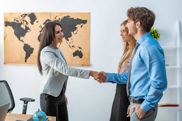 Smiling travel agent shaking hands with happy man standing near girlfriend — Stock Photo