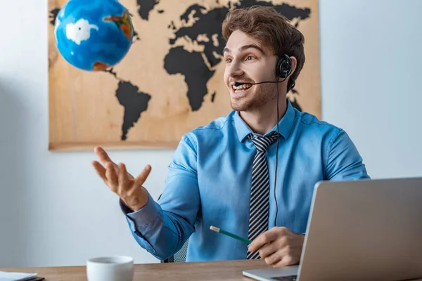 Cheerful travel agent throwing up globe while sitting at workplace — Stock Photo