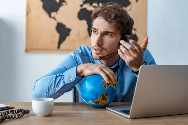 Agente de viajes serio tocar globo mientras trabaja cerca de la computadora portátil - foto de stock
