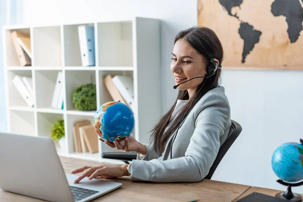 Agent de voyage positif tenant globe tout en étant assis sur le lieu de travail — Photo de stock