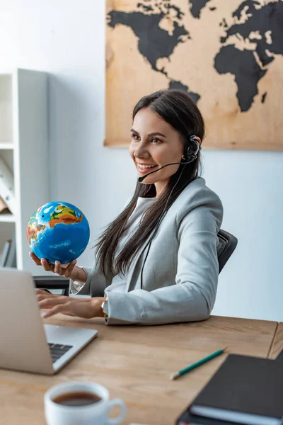 Smiling travel agent holding globe while sitting at workplace — Stock Photo