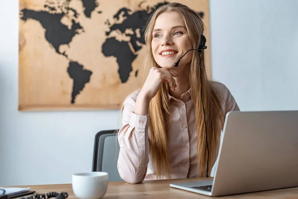 Sorrindo agente de viagens olhando para longe enquanto sentado no local de trabalho — Fotografia de Stock