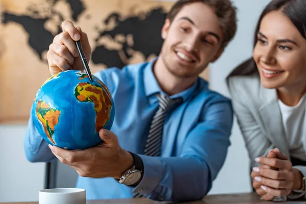 Selective focus of attractive travel agent standing near smiling colleague pointing with pen at globe — Stock Photo