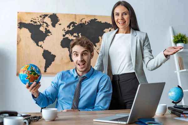 Attractive travel agent showing welcome gesture while standing near smiling colleague holding globe — Stock Photo