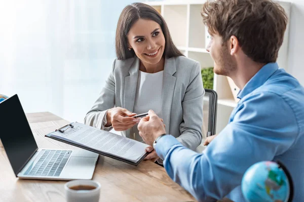 Smiling travel agent giving contract and pen to young client — Stock Photo