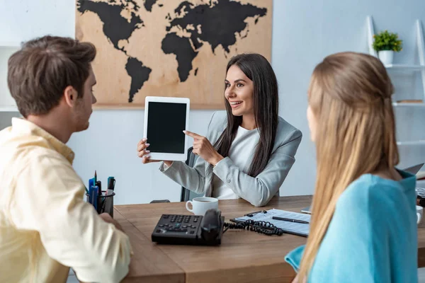 Lächelndes Reisebüro zeigt mit Finger und digitalem Tablet in die Nähe einiger Touristen — Stockfoto