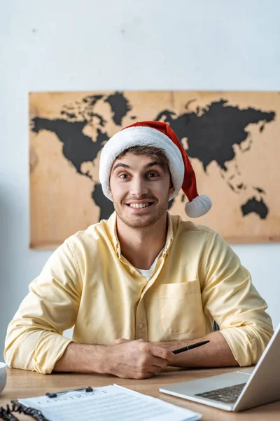 Веселий туристичний агент в капелюсі Санта дивиться на камеру, сидячи на робочому місці — стокове фото