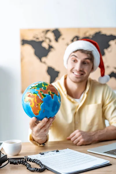 Selective focus of smiling travel agent in santa hat holding globe while sitting at workplace — Stock Photo
