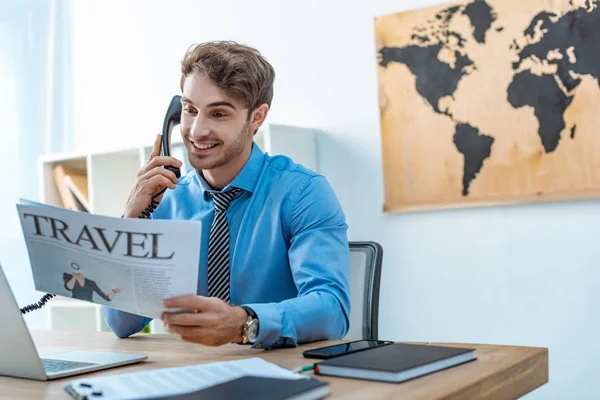 Agente de viajes sonriente hablando por teléfono mientras sostiene el periódico de viaje - foto de stock