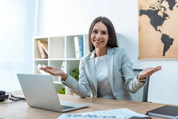 Attractive travel agent smiling at camera and showing welcome gesture while sitting at workplace — Stock Photo