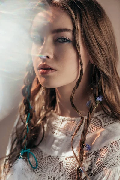 Atractiva chica étnica con trenzas en peinado posando en vestido boho blanco sobre gris con bengalas de lente - foto de stock