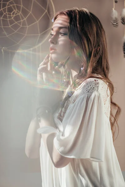 Elegante chica con trenzas en peinado posando en vestido boho blanco sobre gris con bengalas de lente - foto de stock