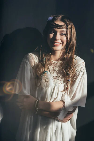 Atractiva chica feliz con trenzas en peinado posando en vestido boho blanco sobre gris con bengalas de lente - foto de stock