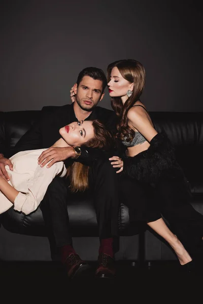 Attraktive und stilvolle Frauen liegen und umarmen gutaussehenden Mann isoliert auf schwarz — Stockfoto