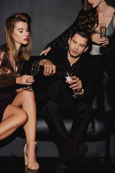 Красивый мужчина разливает шампанское для привлекательных и стильных женщин, изолированных на черном — стоковое фото