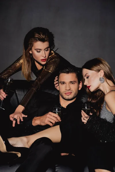 Mujeres atractivas y hombre guapo sosteniendo copas de champán aisladas en negro - foto de stock